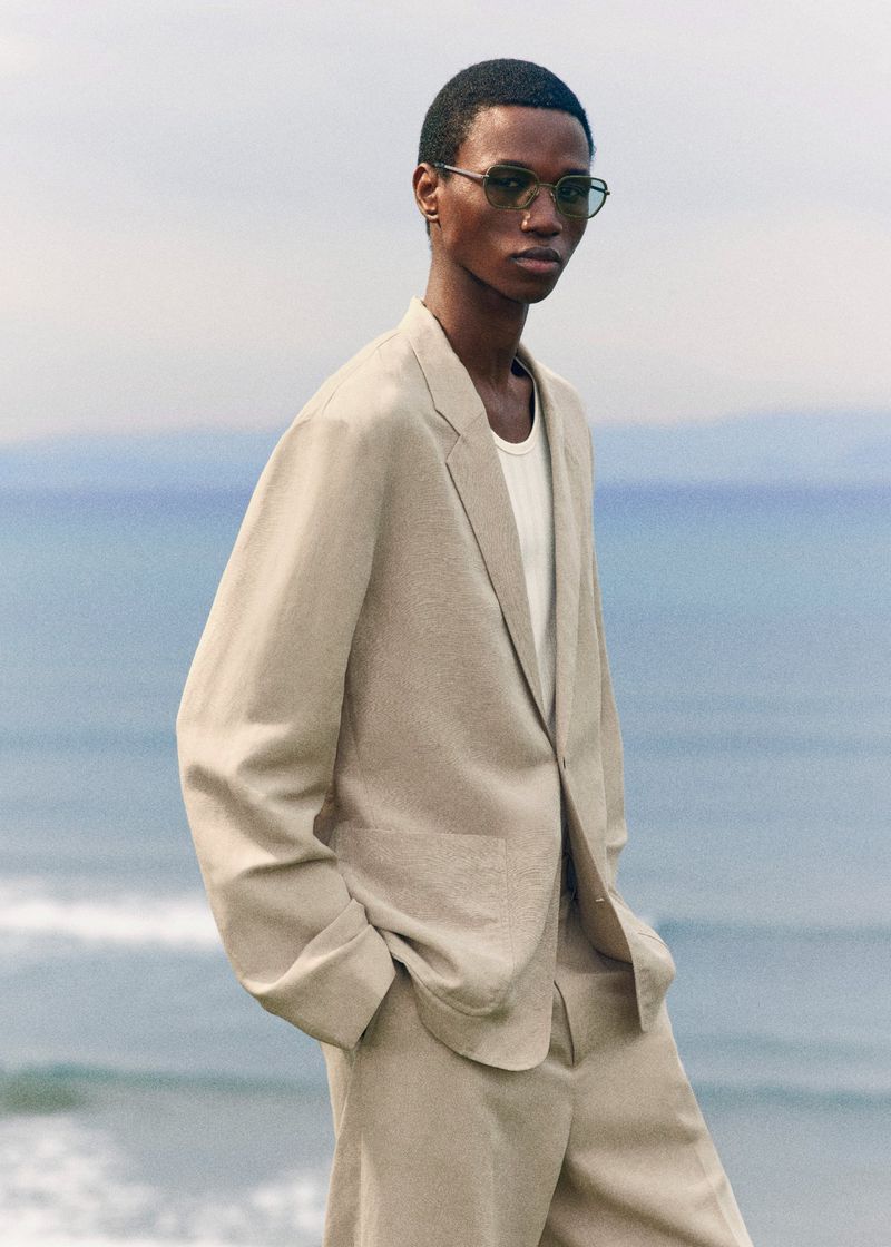 Model Gaye Serigne poses wearing a relaxed beige suit jacket and tinted sunglasses against an ocean backdrop for COS.