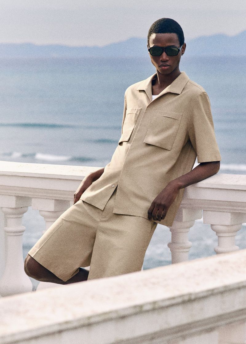Model Gaye Serigne wearing a beige short-sleeve shirt, matching shorts, and dark sunglasses, leaning on a white balustrade by the ocean.