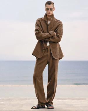 Model wearing a brown tailored jacket, matching wide-leg trousers, sunglasses, and black sandals against a calm sea background.