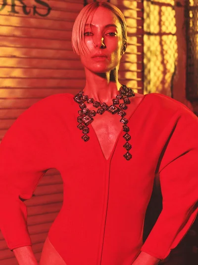 Model Jessica Miller wearing a red sculptural bodysuit and geometric statement necklace illuminated under warm amber light.