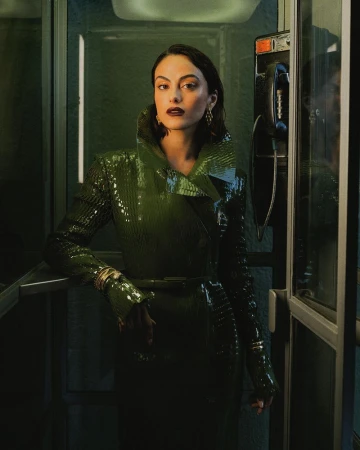 Camila Mendes wearing an embossed green patent coat inside a glass telephone booth with a vintage payphone.
