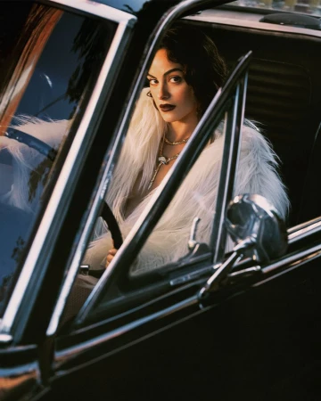 Camila Mendes wearing a voluminous white faux fur coat and necklace while seated inside a black vintage car.