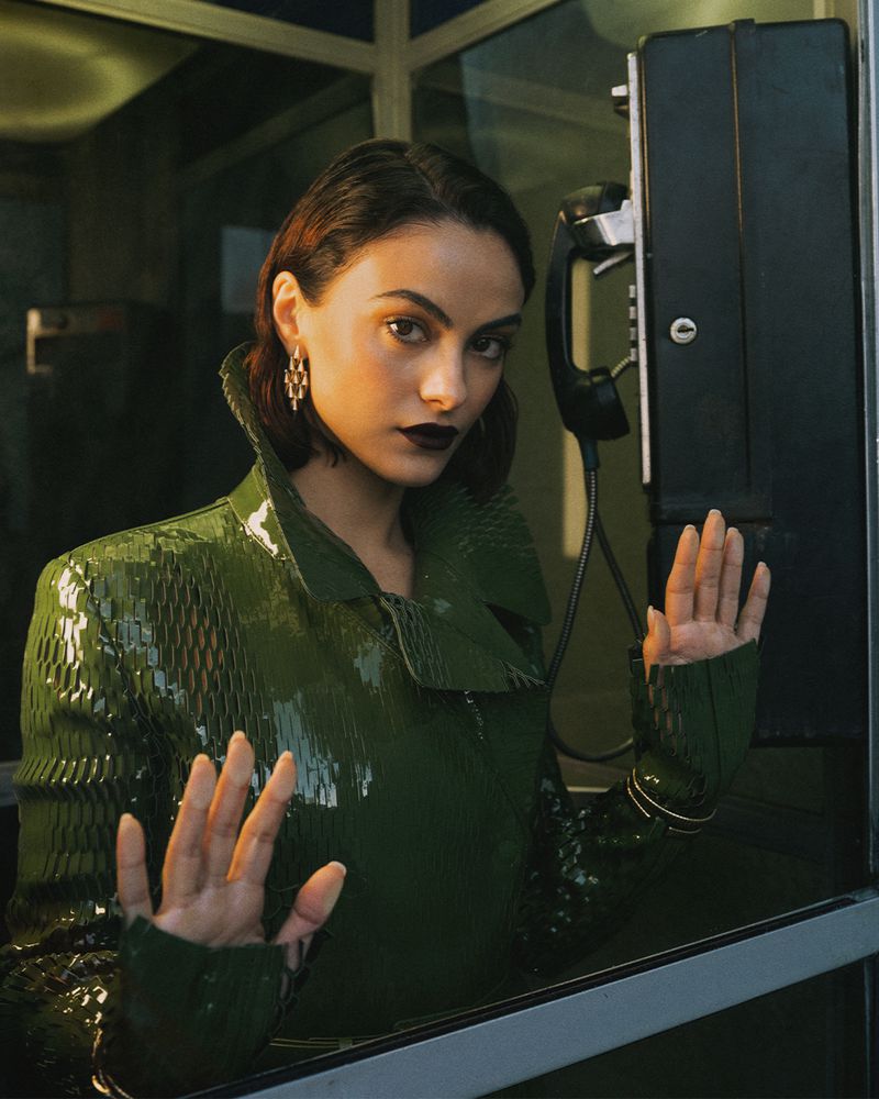 Camila Mendes wearing a glossy olive green textured jacket with her hands pressed against a phone booth glass window.