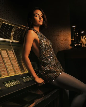 Camila Mendes wearing a sequined top and stockings seated beside a vintage jukebox in a dimly lit bar setting.