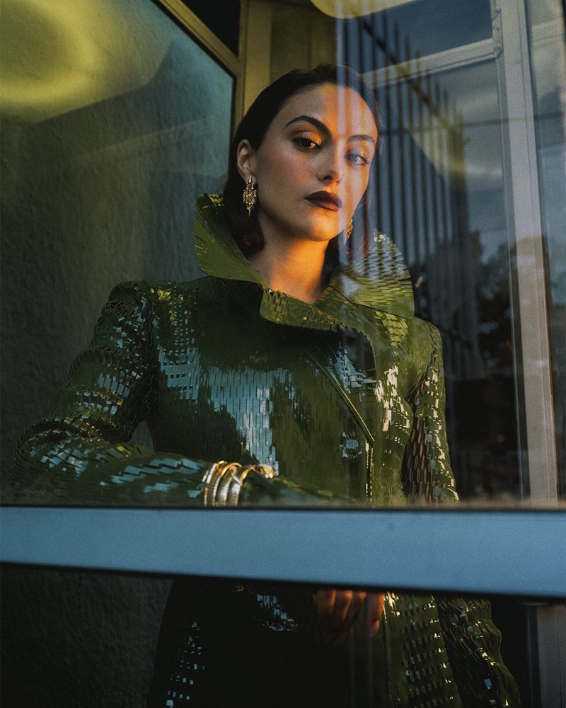 Camila Mendes wearing a high-collared glossy green coat and gold earrings viewed through a glass window reflection.