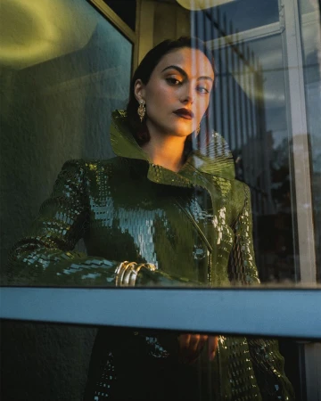 Camila Mendes wearing a high-collared glossy green coat and gold earrings viewed through a glass window reflection.