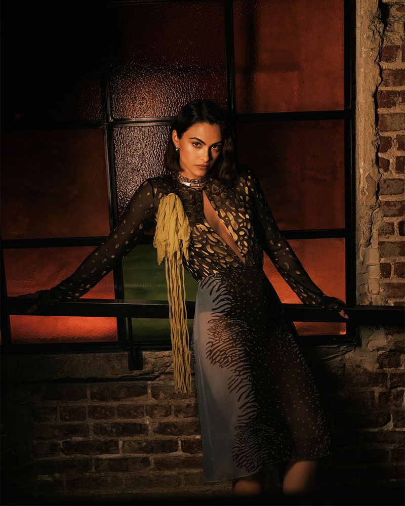 Camila Mendes wearing patterned black dress leaning against dark window and brick wall