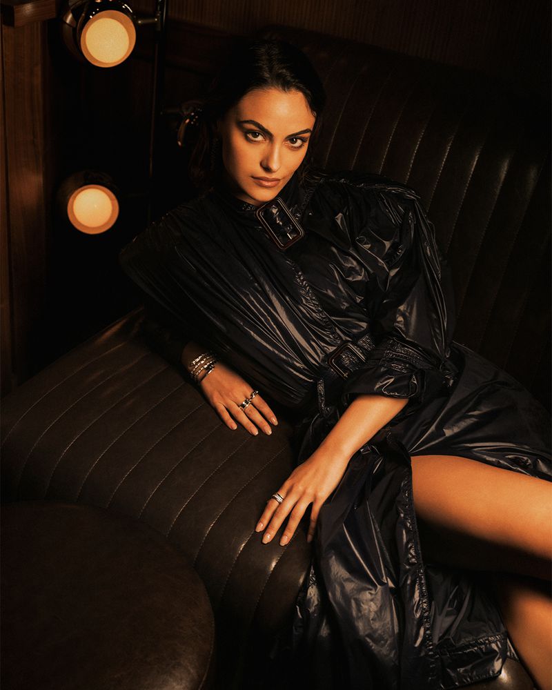 Camila Mendes wearing a glossy dark belted trench coat while seated in a dark leather booth illuminated by warm wall lights.