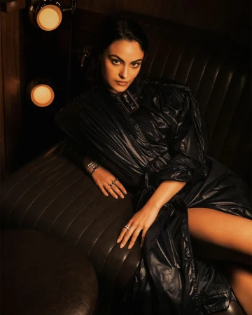 Camila Mendes wearing a glossy dark belted trench coat while seated in a dark leather booth illuminated by warm wall lights.