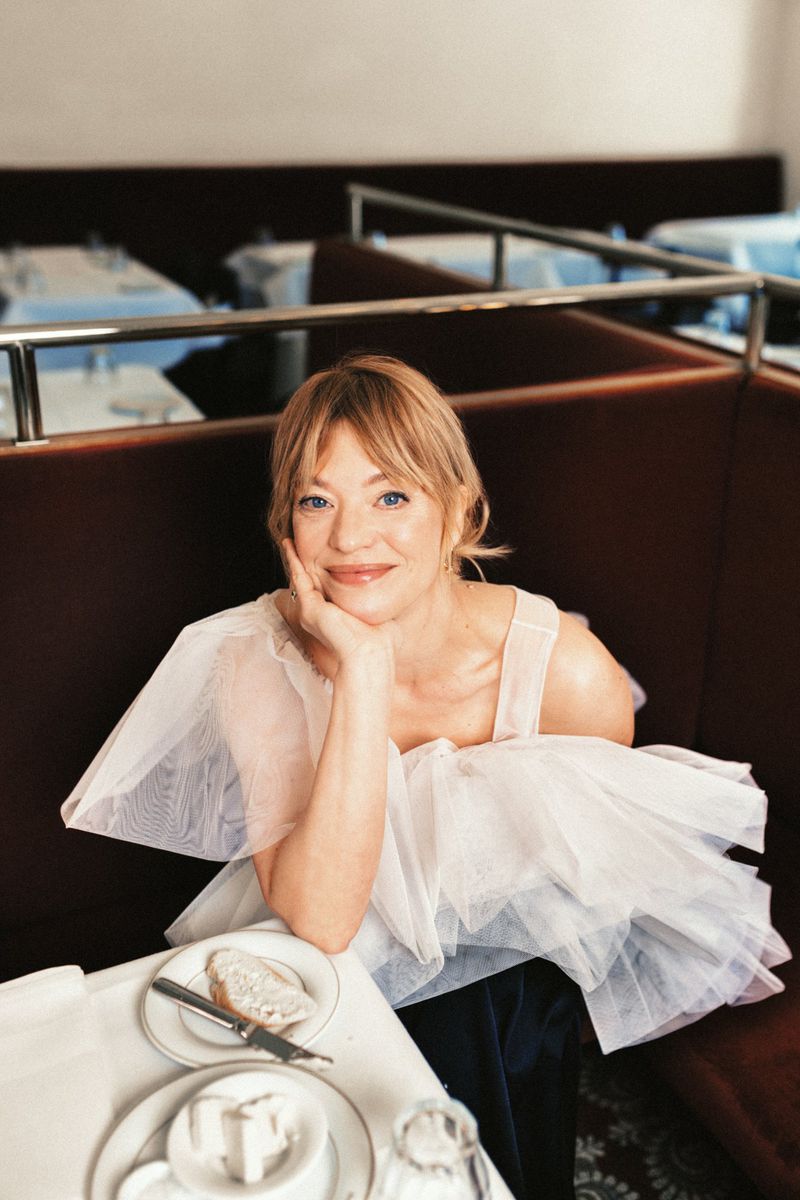 Blonde woman in a voluminous white tulle top resting her chin on her hand while seated in a dark restaurant booth.