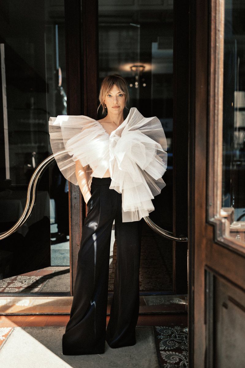 Woman wearing a sculptural white ruffled tulle top and wide-leg black trousers standing in an open doorway.