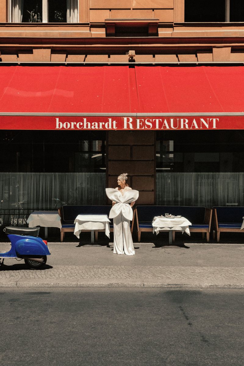 Model in a sculptural white jumpsuit posing in front of Borchardt restaurant under a bright red awning with a blue scooter.