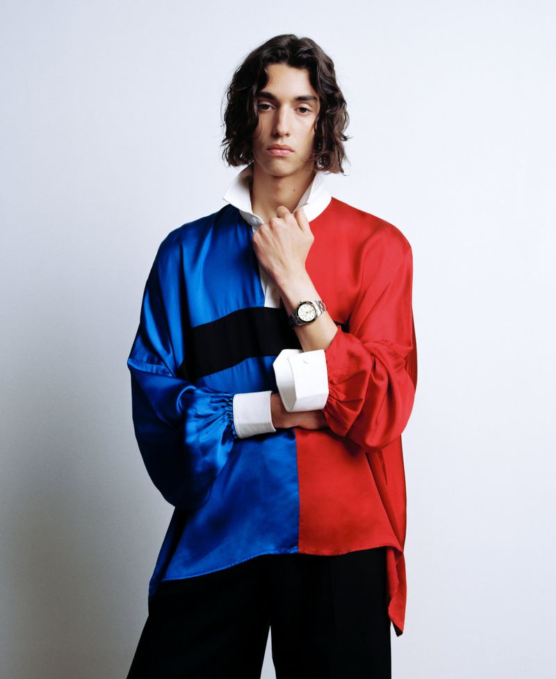 Model with long dark hair wearing a split-color blue and red shirt with white cuffs against a light background.