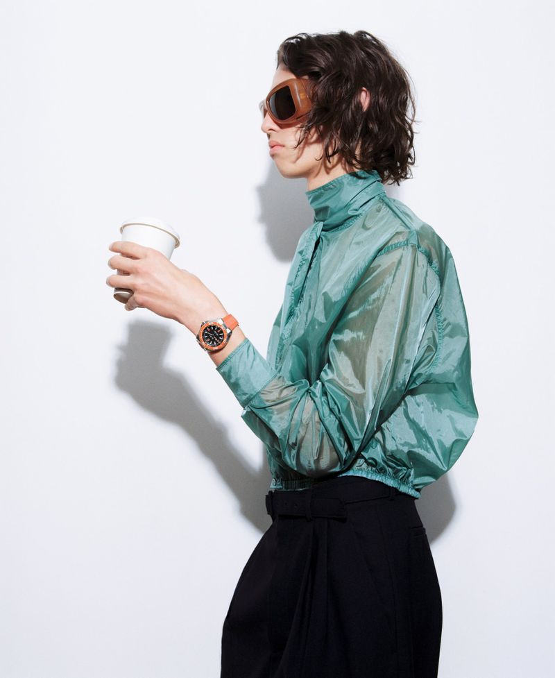 Model in profile wearing oversized sunglasses, a translucent teal jacket, black trousers, and an orange watch, holding a paper coffee cup.
