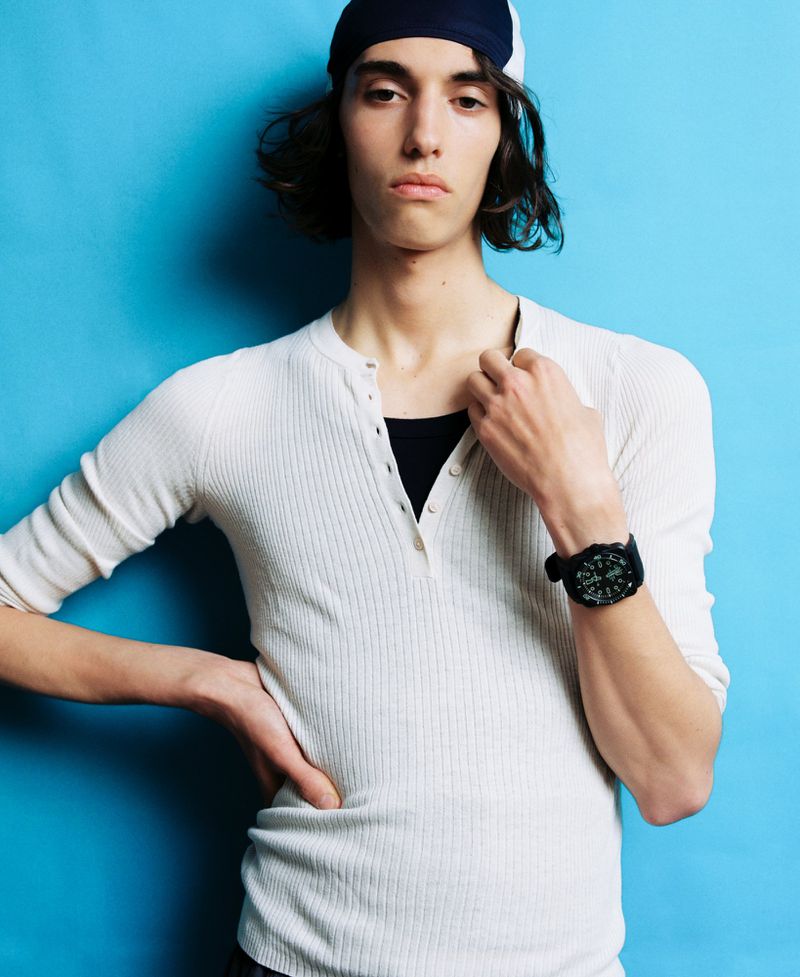 Model Nour Oitahar Normand wearing a white henley shirt and dark head covering against a bright blue background