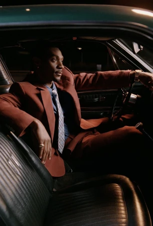 Jonathan Daviss seated in the driver's seat of a vintage green car wearing a rust brown suit and patterned tie at night.