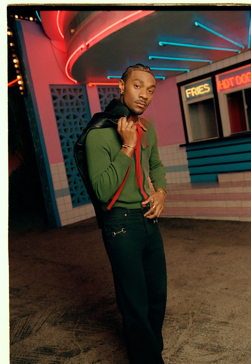 Jonathan Daviss wearing a green sweater and dark trousers holding a black jacket outside a neon-lit diner at night.