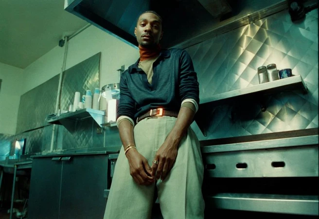 Portrait of a Black man leaning against stainless steel counters in a commercial kitchen setting.