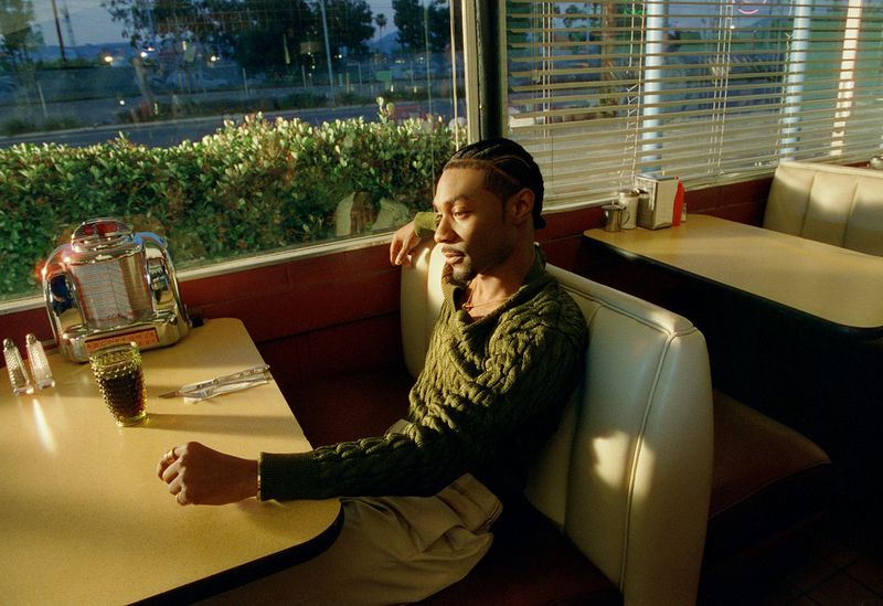 Man seated in a booth at a retro diner wearing a green cable-knit sweater, with a vintage tabletop jukebox beside him.
