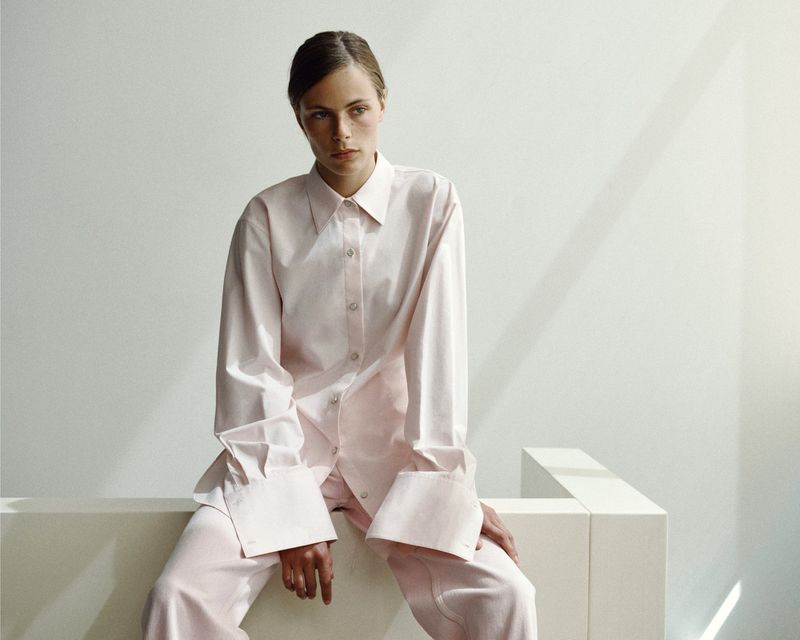 Model wearing an oversized pale pink button-down shirt and trousers seated on a white block against a neutral studio wall.