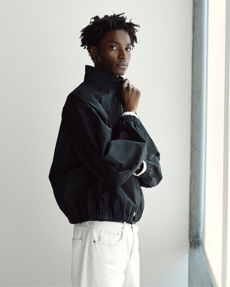 A Black male model posing in a black high-collar zip jacket and white jeans against a bright minimalist studio wall.