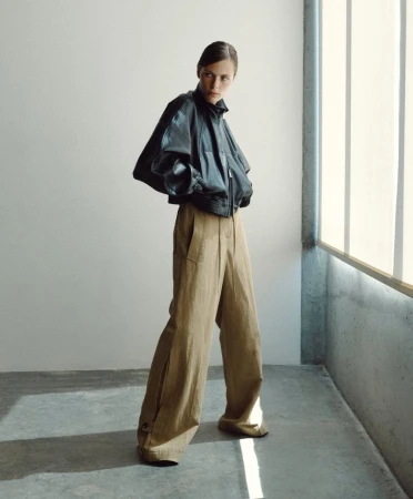 Model wearing an oversized black leather jacket and wide-leg tan trousers standing near a window on a concrete floor.