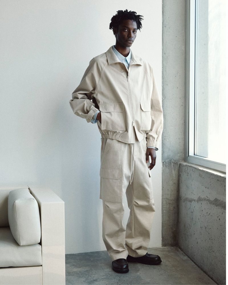 Model wearing a neutral beige jacket and matching trousers standing near a large window in a modern minimal interior.
