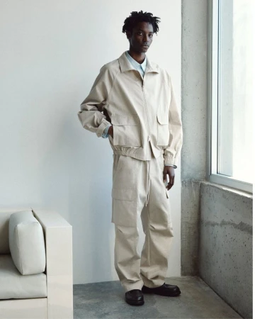 Model wearing a neutral beige jacket and matching trousers standing near a large window in a modern minimal interior.