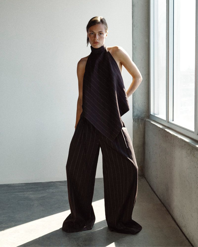 Model wearing a dark pinstripe halter top and matching oversized wide-leg trousers standing near an industrial window.
