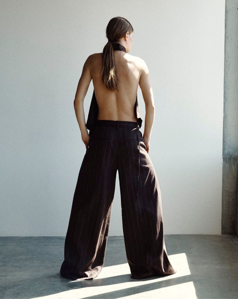 Model seen from behind wearing an open-back top and dark wide-leg pinstripe trousers in a sunlit minimalist studio.
