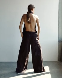 Model seen from behind wearing an open-back top and dark wide-leg pinstripe trousers in a sunlit minimalist studio.