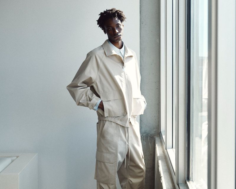 A Black man standing by a large window wearing a light beige zip-up jacket and matching cargo pants.