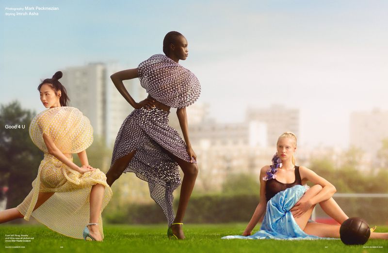 Three fashion models posing in sculptural couture garments on a green lawn with city buildings in the background under warm daylight.