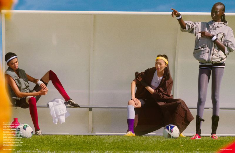 Three models styled in athletic sportswear pose on a soccer pitch dugout bench alongside footballs on the grass under bright sunlight.