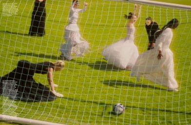 Models in voluminous black and white evening dresses play soccer on a sunny grass pitch viewed through a white goal net.