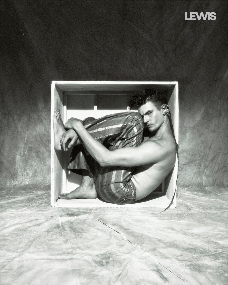 Model in striped pants curled inside a small white box against a textured canvas backdrop.