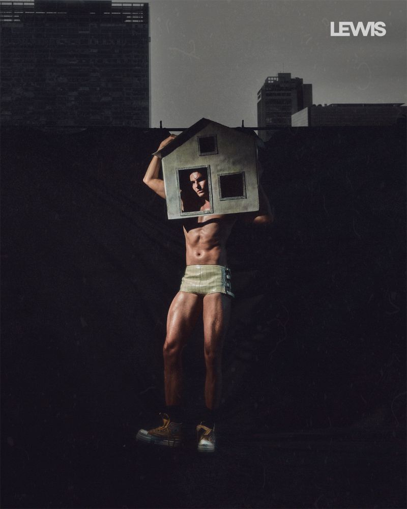 Muscular man wearing shorts and high-top shoes with a miniature model house over his head, photographed against a dark city backdrop.
