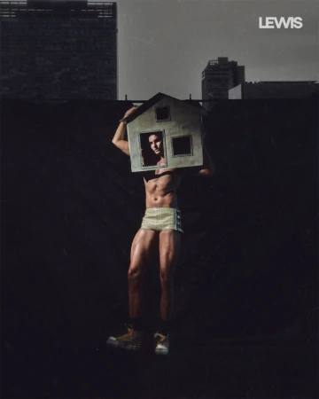 Muscular man wearing shorts and high-top shoes with a miniature model house over his head, photographed against a dark city backdrop.