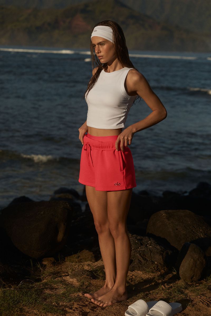 Woman in white top and coral shorts on a rocky beach with ocean and hills in the background, accessorized with a white headband.