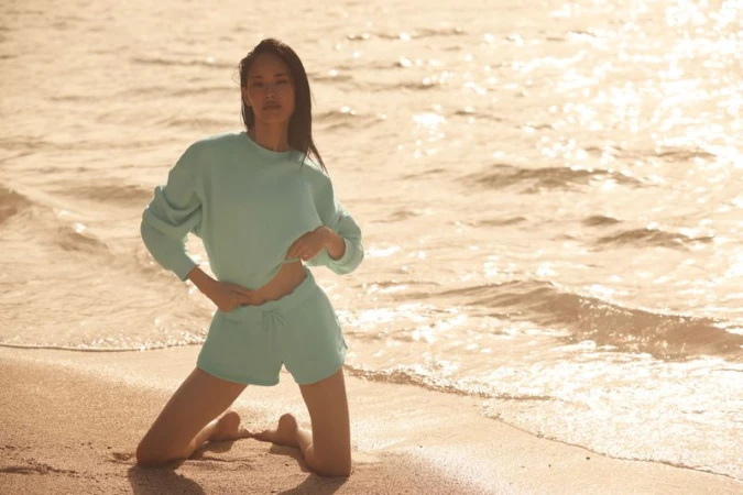 A woman kneels on the beach in light blue matching lounge wear, a cropped sweater and shorts with the ocean and sunlight background.