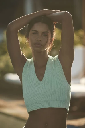 Portrait of a woman with arms raised behind her head, wearing a light green crop top, posing outdoors in natural light.