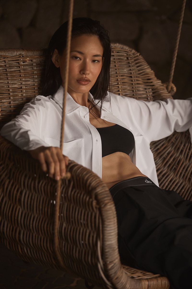 Portrait of an Asian adult female in a white shirt and black bra, sitting in a wicker chair with a stone wall background.