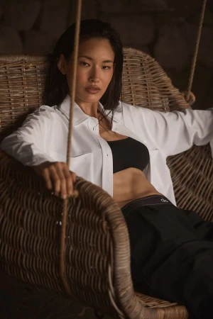 Portrait of an Asian adult female in a white shirt and black bra, sitting in a wicker chair with a stone wall background.