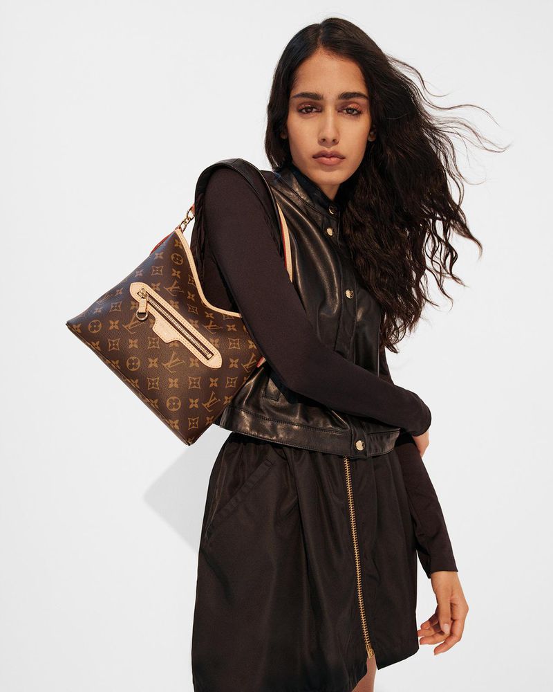 Model with long dark hair wearing black leather vest and holding monogram handbag against white background