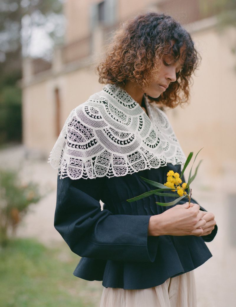 Woman with curly hair wearing an intricate white lace collar over a navy peplum jacket while holding a sprig of yellow flowers outdoors.