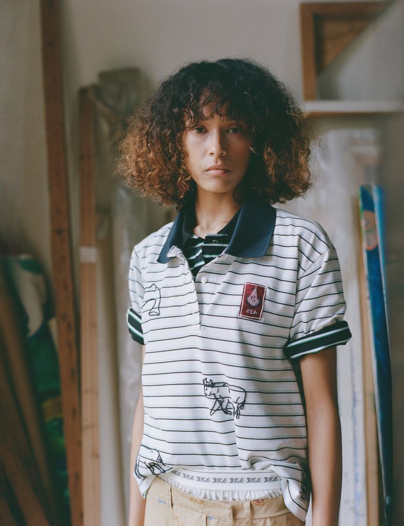 Model with curly ombre hair wearing a white striped polo shirt standing in an art studio for Sea PF26 Editorial.
