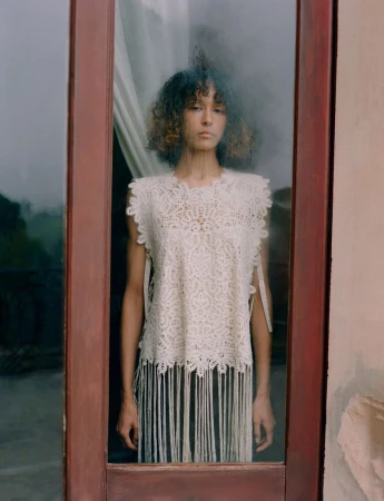 Model with curly hair wearing an off-white lace crochet top with fringe detailing, seen looking through a glass door pane.