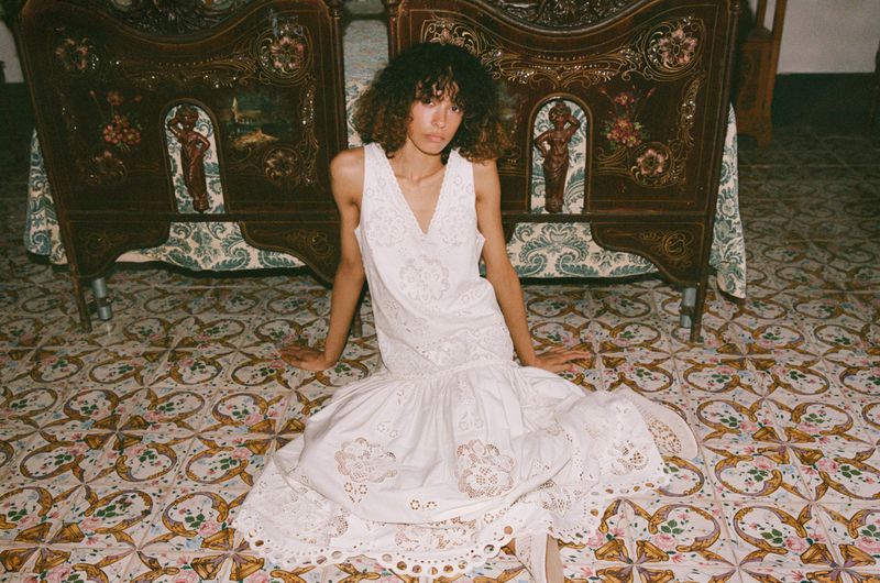 Model with dark curly hair wearing a white embroidered dress seated on patterned tile floor before ornate wooden headboards.