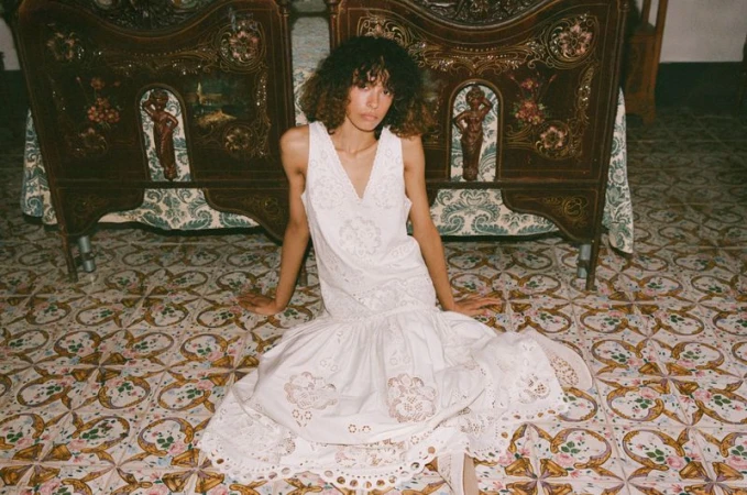 Model with dark curly hair wearing a white embroidered dress seated on patterned tile floor before ornate wooden headboards.