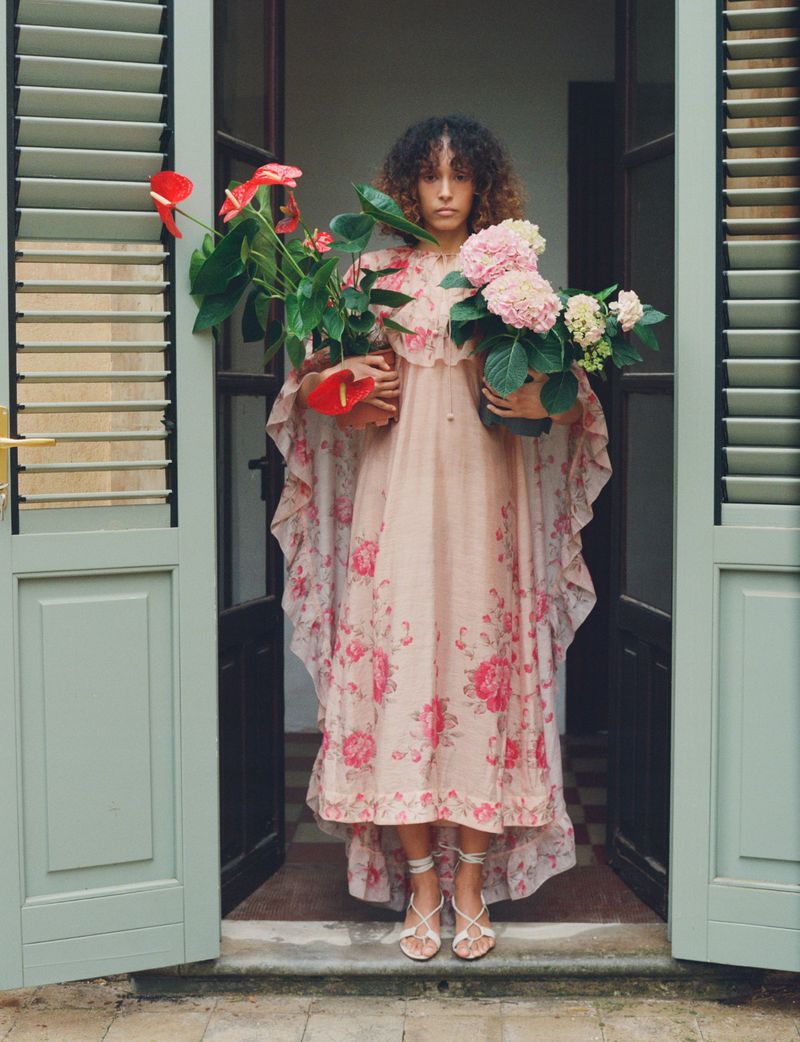 Model wearing a pink floral cape-sleeve maxi dress and holding potted anthurium and hydrangea plants in an open doorway.